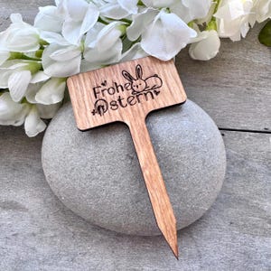 May include: A wooden Easter garden stake with the words "Frohe Ostern" and a bunny illustration. The stake is light brown and has a pointed end for easy insertion into the ground. The stake is resting on a gray stone.