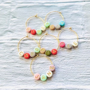May include: Five gold-toned wine glass charms with colorful beads spelling out the names "BINEVER", "CAROPIA", "JAINE", "FEE", and "VERA".