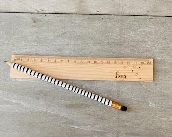 May include: A wooden ruler with a black and white striped pencil lying on top of it. The ruler is marked with centimetres and has the name "Fiona" engraved on it with stars.