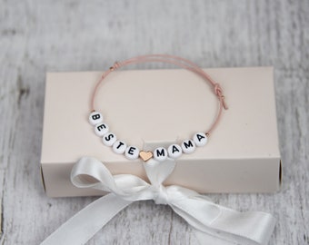 May include: A white bracelet with the words "BESTE MAMA" spelled out in black letters on white beads. A small gold heart charm is between the words.