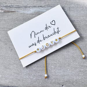 Könnte beinhalten: Eine weiße Karte mit dem Text "Nimm dir was du brauchst" und einem Herzsymbol. Ein goldfarbenes Schnurarmband mit weißen Perlen, die "JOY" buchstabieren, ist an der Karte befestigt.