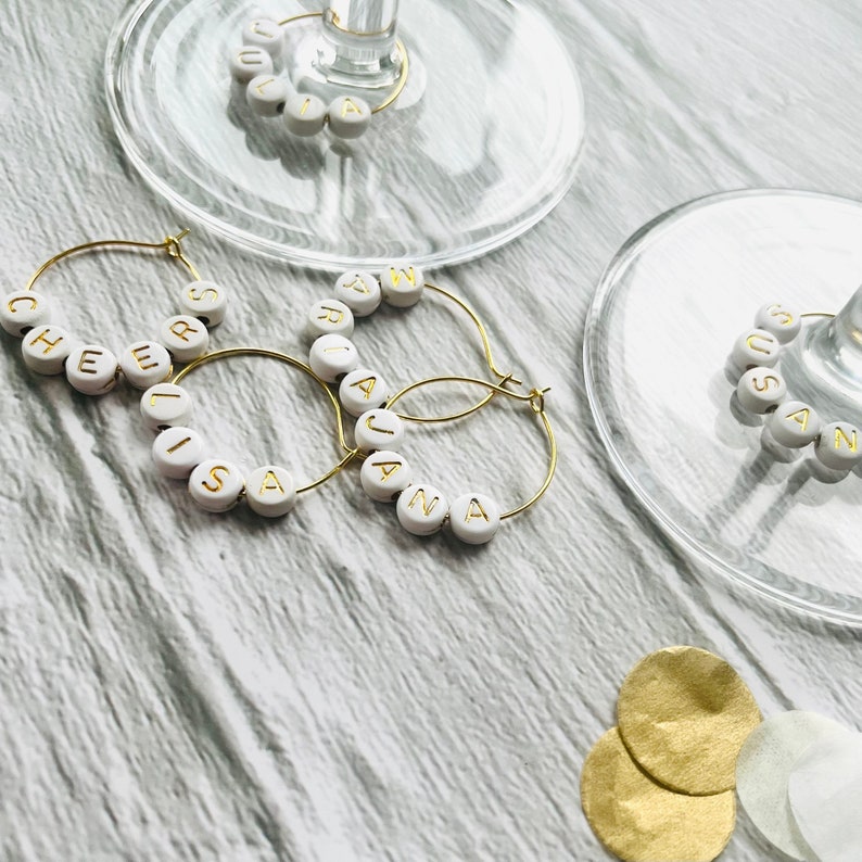 May include: Four gold-toned hoop-shaped glass charms with white beads spelling out the names "CHEERS", "LISA", "ARIANA", and "SUSAN".