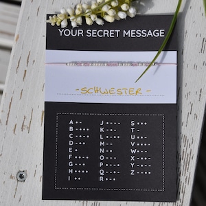 Peut inclure: Une carte noire avec le texte "YOUR SECRET MESSAGE" et "SCHWESTER" en lettres dorées. La carte comprend également un tableau avec l'alphabet et les symboles de code Morse correspondants.