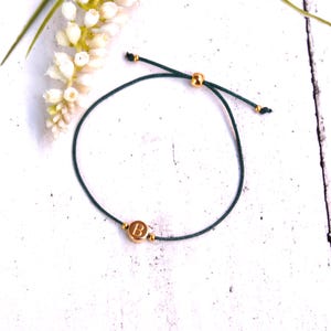 Könnte beinhalten: Ein dunkelgrünes Kordelarmband mit einer einzelnen runden goldfarbenen Perle, die mit dem Buchstaben B graviert ist. Das Armband verfügt über einen verschiebbaren goldfarbenen Verschluss und zwei kleine goldfarbene Abstandsperlen.