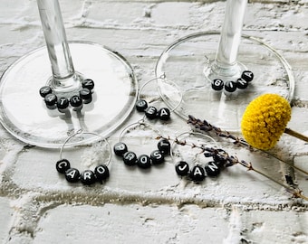 May include: Five black beaded wine glass charms with the names "CARO", "PROST", "BEA", "GINA", and "JAN" spelled out on them. The charms are on a white brick background.