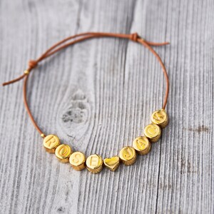 以下が含まれることがあります： ゴールドのビーズで「Rose」と「Jack」という名前を綴った、ブラウンの革紐のブレスレット。ゴールドのハートチャームで区切られています。