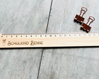 May include: A wooden ruler with a black and white illustration of pencils in a cup. The ruler is marked with numbers from 0 to 20. The text "SCHULKIND BENNE" is engraved on the ruler.