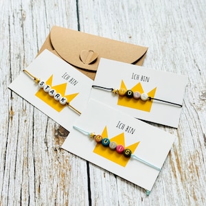 Peut inclure: Trois cartes avec des bracelets en perles colorées. Chaque carte a une couronne jaune avec les mots "Ich bin" et un mot différent sur chaque carte : "stark", "toll" et "mutig".