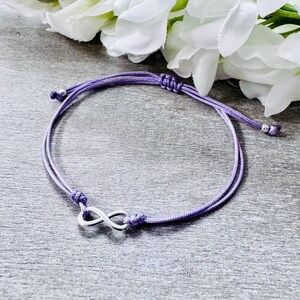Puede incluir: Una pulsera ajustable morada con un símbolo de infinito plateado. La pulsera está hecha de un cordón fino y tejido con nudos corredizos para ajustarla. El dije de infinito está centrado en la pulsera.