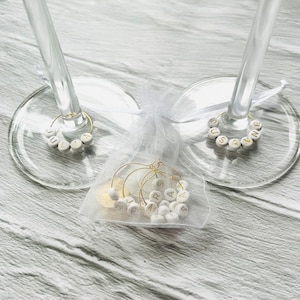 May include: Two wine glass charms with the names "Julia" and "Susanne" spelled out in gold letters on white beads. A white organza bag contains additional charms with gold letters.