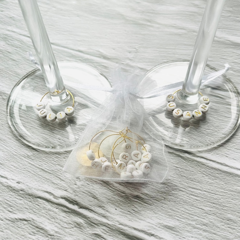 May include: Two wine glass charms with the names "Julia" and "Susanne" spelled out in gold letters on white beads. A white organza bag contains additional charms with gold letters.