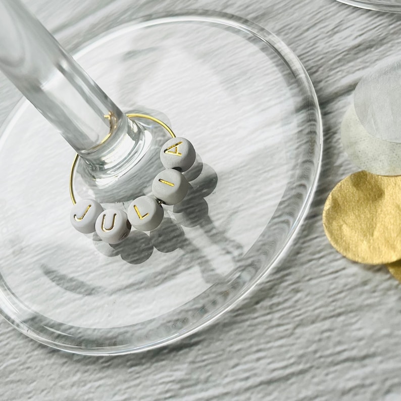 May include: A close-up of a glass wine glass with a gold wire charm that spells out the name "JULIA" in white beads.