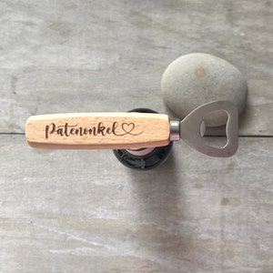 Pode incluir: Um abridor de garrafas de madeira com a palavra "Patenonkel" e um símbolo de coração gravados na pega. O abridor tem uma cabeça de metal prateado e está apoiado numa garrafa escura.