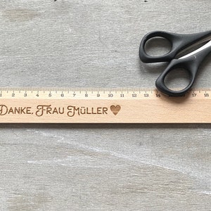 K&ouml;nnte beinhalten: Ein Holzlineal mit einer schwarz-wei&szlig;en Zahlenleiter von 0 bis 20. Das Lineal tr&auml;gt den Text "DANKE, FRAU M&Uuml;LLER" und ein Herzsymbol, das eingraviert ist.