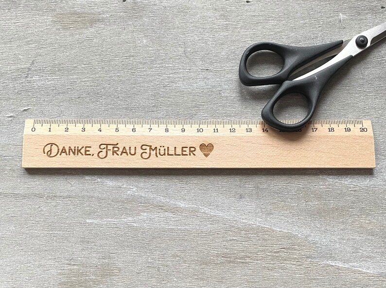 K&ouml;nnte beinhalten: Ein Holzlineal mit einer schwarz-wei&szlig;en Zahlenleiter von 0 bis 20. Das Lineal tr&auml;gt den Text "DANKE, FRAU M&Uuml;LLER" und ein Herzsymbol, das eingraviert ist.