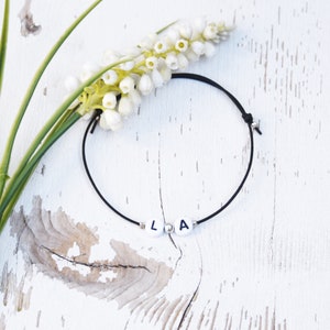 Puede incluir: Un brazalete de cuerda negro con cuentas blancas que deletrean las letras "LA".