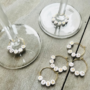 May include: Six gold-colored wine glass charms with white beads spelling out the names "Lisa", "Cheers", "Jana", "Maria", "Julia", and "Susan".