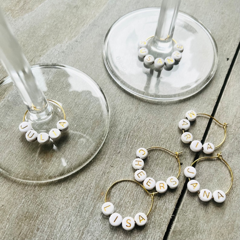 May include: Six gold-colored wine glass charms with white beads spelling out the names "Lisa", "Cheers", "Jana", "Maria", "Julia", and "Susan".