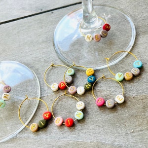 May include: Six gold-toned wine glass charms with colorful beads spelling out the names "Nele", "Vera", "Fee", "Carolin", "Diana", and "Piabin".