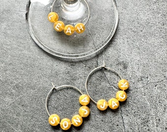 May include: Two gold-colored wine glass charms with white letters spelling out the names "LEAH" and "JACK". A third charm with the name "SONJA" is also pictured.