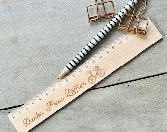 May include: A wooden ruler with a black and white striped pencil, two gold binder clips, and the text "Danke. Frau Löffler" engraved on the ruler.