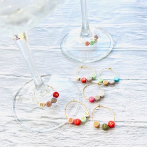 May include: Five gold-colored wine glass charms with colorful beads spelling out the names "Nele", "Fefe", "Bine", "Pia", and "Jara".