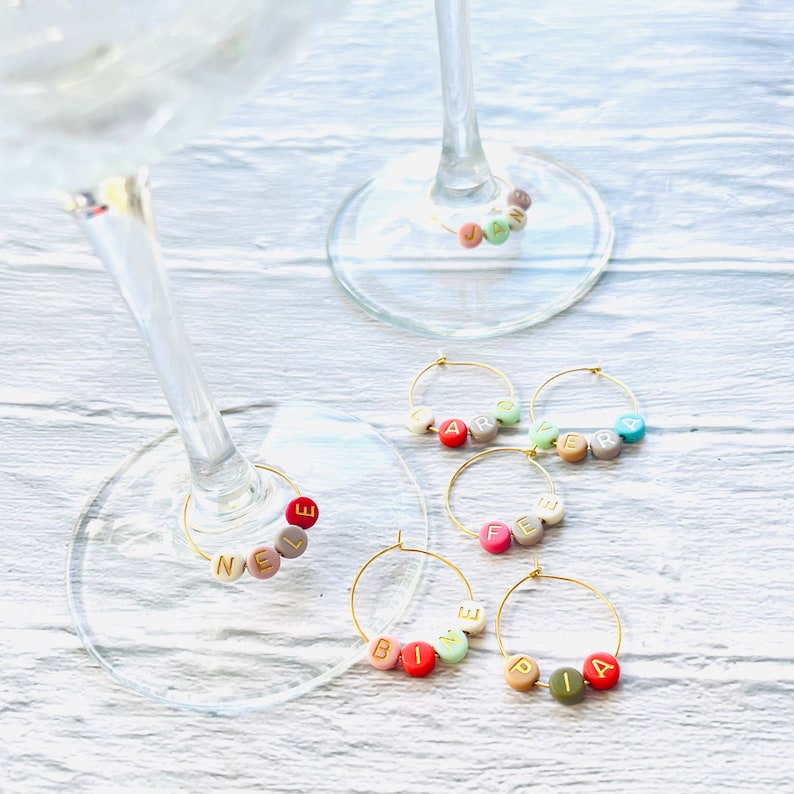 May include: Five gold-colored wine glass charms with colorful beads spelling out the names "Nele", "Fefe", "Bine", "Pia", and "Jara".