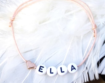 May include: A bracelet featuring a thin, light pink cord with adjustable knots. The bracelet includes four white, round beads with the black letters E, L, L, and A printed on them, accompanied by a small, metallic, rose gold-coloured heart charm.