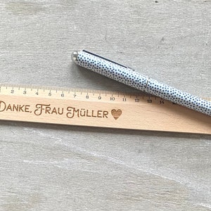 K&ouml;nnte beinhalten: Ein Holzlineal mit einem Herz und dem eingravierten Text "DANKE, FRAU M&Uuml;LLER". Ein wei&szlig;er Stift mit schwarzen Punkten liegt auf dem Lineal.