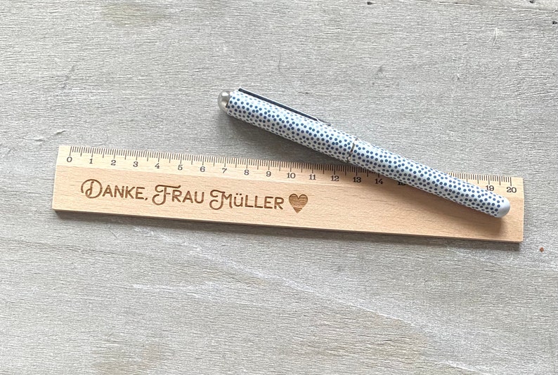 K&ouml;nnte beinhalten: Ein Holzlineal mit einem Herz und dem eingravierten Text "DANKE, FRAU M&Uuml;LLER". Ein wei&szlig;er Stift mit schwarzen Punkten liegt auf dem Lineal.