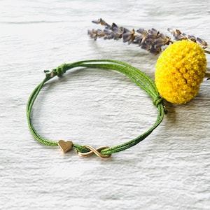 Puede incluir: Un brazalete de cuerda verde con un símbolo de infinito dorado y un pequeño dije en forma de corazón.