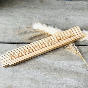 Könnte beinhalten: Ein Holz-Klapplineal mit den Namen "Kathrin & Paul" und einem kleinen Haussymbol.
