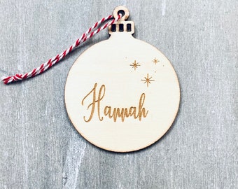 May include: A wooden Christmas bauble ornament with the name "Hannah" engraved on it. The ornament has a red and white twine string attached to the top.