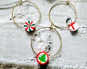 May include: Three gold hoop earrings with Christmas-themed charms. The charms are a red and white candy cane, a white snowman with a red scarf, and a red Christmas tree with green and yellow details.