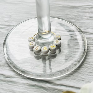 May include: White glass wine charm with the name "SUSANNE" spelled out in gold letters.