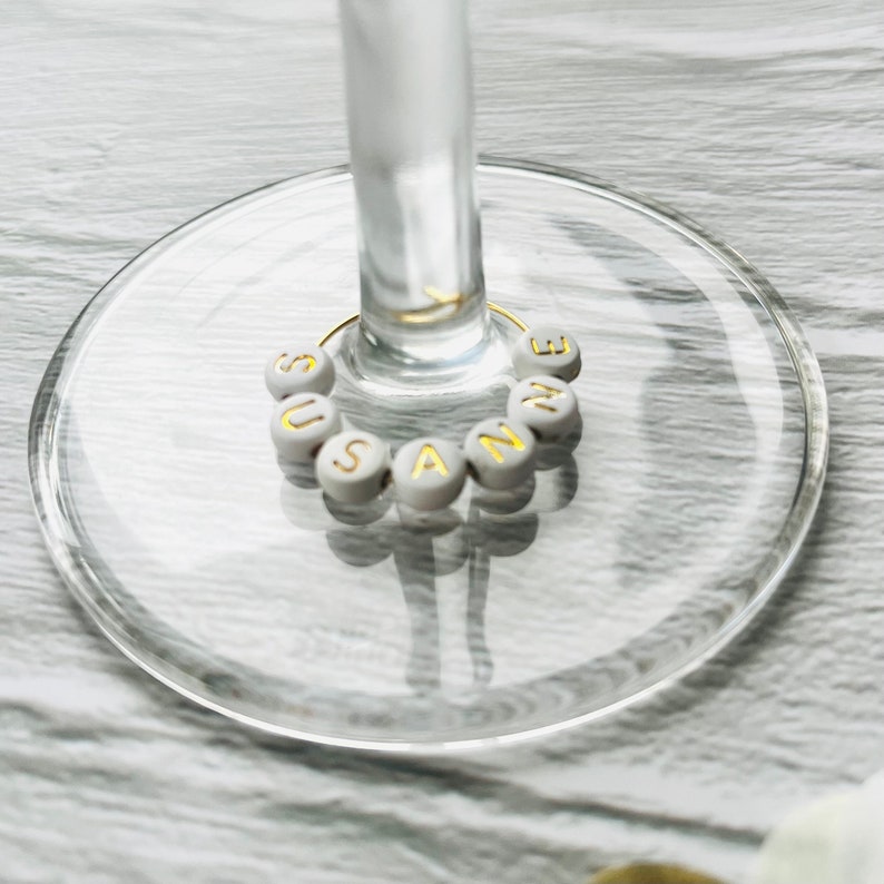 May include: White glass wine charm with the name "SUSANNE" spelled out in gold letters.