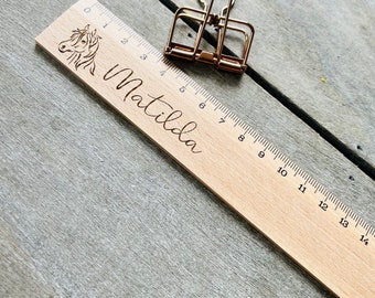 May include: A wooden ruler with engraved measurements and the name "Matilda" in cursive. A horse head is etched on the left side. A gold-coloured binder clip rests on the ruler. The ruler is approximately 35.5 cm long.