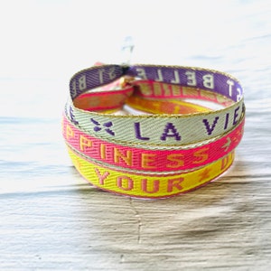 Puede incluir: Pulseras tejidas multicolores con las palabras "Believe", "La Vie", "Happiness" y "Your" en letras amarillas.