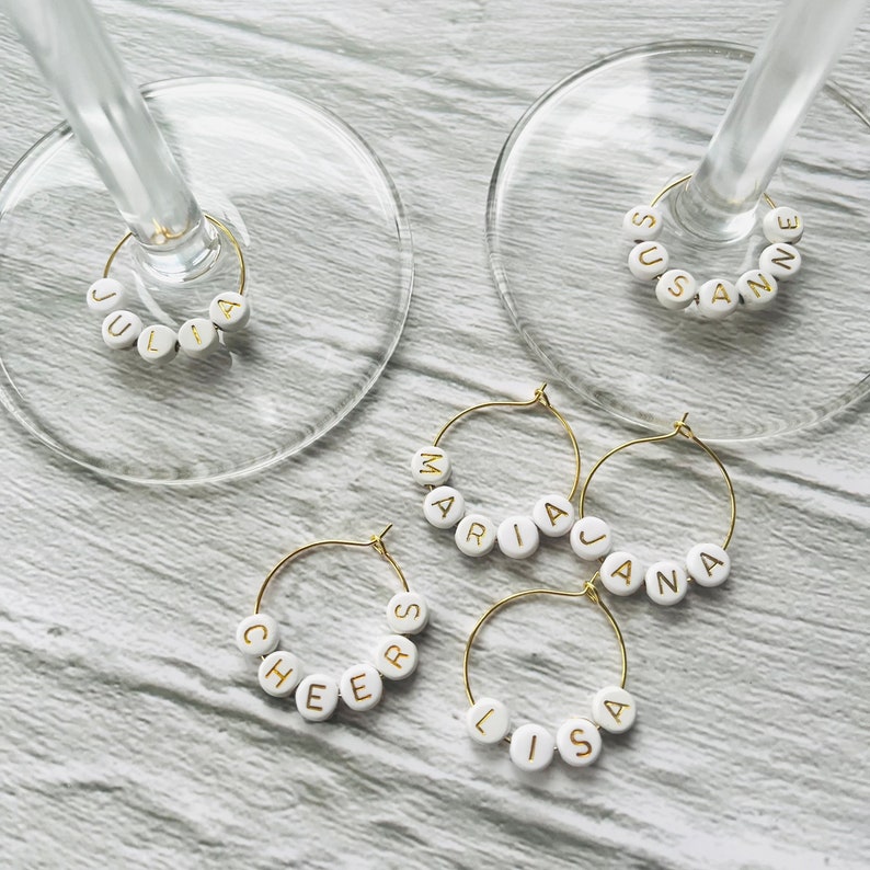 May include: Five gold-colored wine glass charms with white beads spelling out names, including "Julia", "Susan", "Maria", "Jana", and "Lisa".