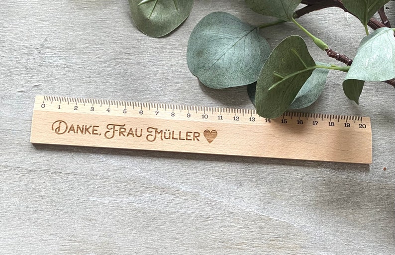 K&ouml;nnte beinhalten: Ein Holzlineal mit einem schwarzen Herz und dem eingravierten Text "DANKE, FRAU M&Uuml;LLER".
