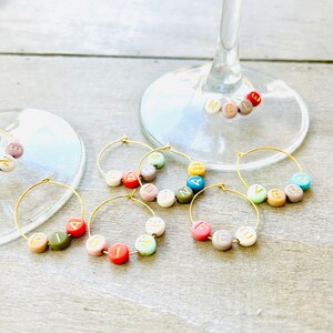 May include: Set of six gold-toned wine glass charms with colorful beads spelling out names. The names are: "ABIGAIL", "FELICIA", "EZRA", "CARDINA", "HAVANA", and "VERA".