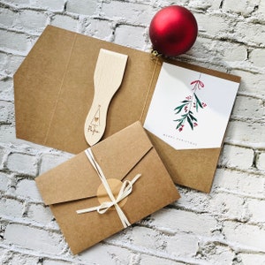May include: A brown paper envelope with a white ribbon and a round sticker. Inside the envelope is a card with a watercolor illustration of a sprig of mistletoe with red berries. The card reads "Merry Christmas".