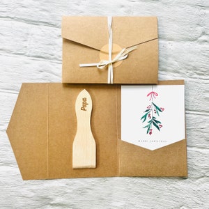 May include: A kraft paper gift set featuring a wooden spatula engraved with "Papa", a Christmas card with a mistletoe illustration, and a matching envelope. The set is tied with a natural twine.