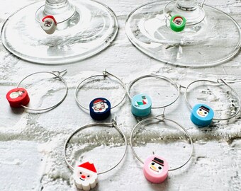 May include: Set of six Christmas-themed wine glass charms. Each charm features a different design, including Santa Claus, a snowman, and a stocking. The charms are made of metal and have a silver finish.