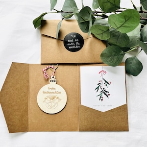 May include: A brown envelope with a black circle sticker that says "meet me under the mistletoe!"  The envelope is open to reveal a card with a hand-drawn illustration of a sprig of mistletoe with red berries.  The card is tucked into a brown envelope with a wooden ornament that says "Frohe Weihnachten" in gold lettering.