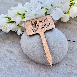 May include: A wooden garden stake with a sign that says "Alles Gute!" and a small watering can with flowers. The stake is resting on a grey rock.