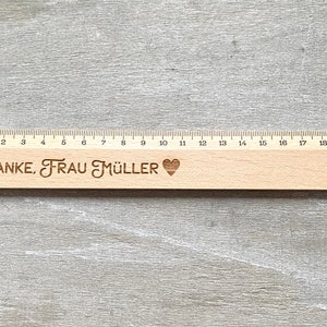 K&ouml;nnte beinhalten: Ein Holzlineal mit einer schwarzen und wei&szlig;en Zahlenleiter von 0 bis 20. Das Lineal tr&auml;gt den Text "DANKE, FRAU M&Uuml;LLER" und ein Herzsymbol, das eingraviert ist.