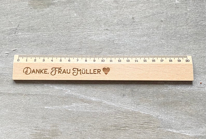 K&ouml;nnte beinhalten: Ein Holzlineal mit einer schwarzen und wei&szlig;en Zahlenleiter von 0 bis 20. Das Lineal tr&auml;gt den Text "DANKE, FRAU M&Uuml;LLER" und ein Herzsymbol, das eingraviert ist.