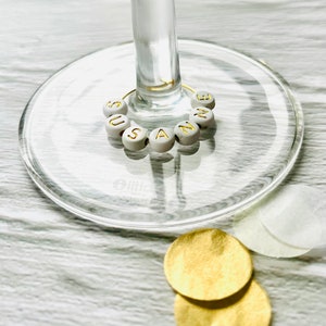 May include: White glass wine glass with a gold wire charm with the name "Suzanne" spelled out in gold letters on white beads.