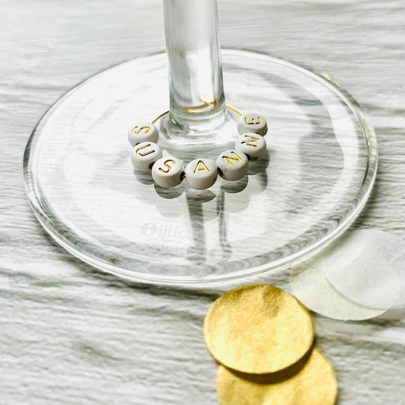 May include: White glass wine glass with a gold wire charm with the name "Suzanne" spelled out in gold letters on white beads.